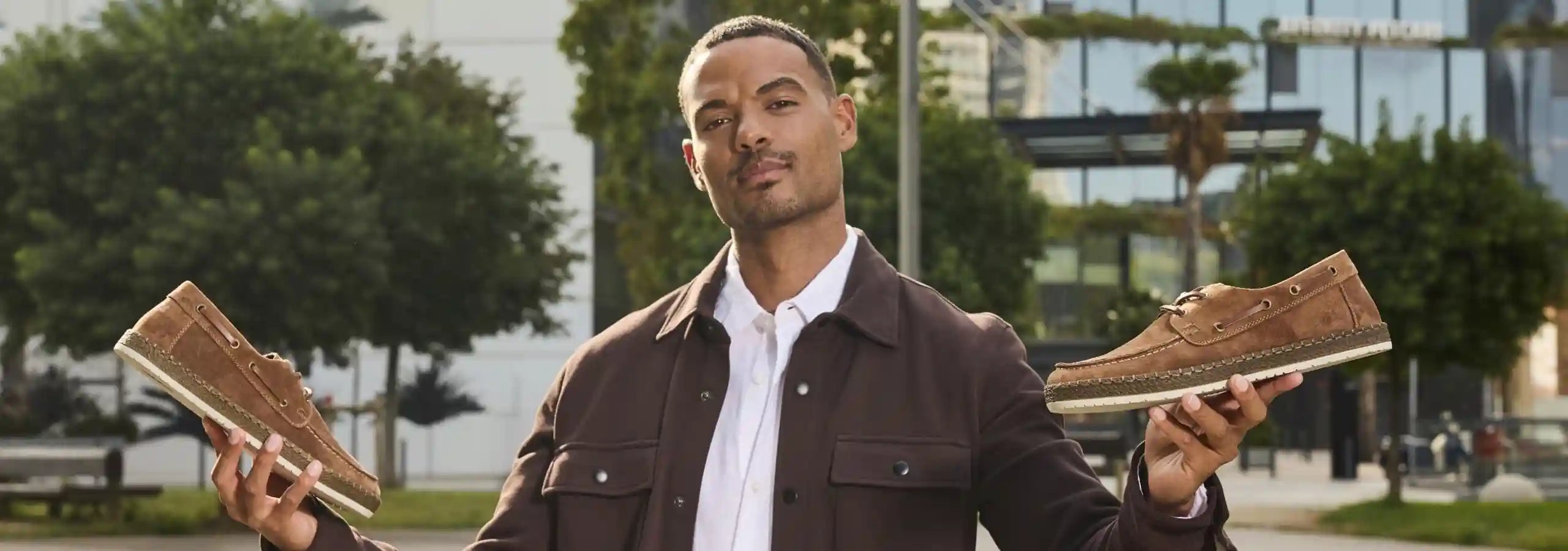 Man holding two pairs of brown shoes outdoors with trees and buildings in the background